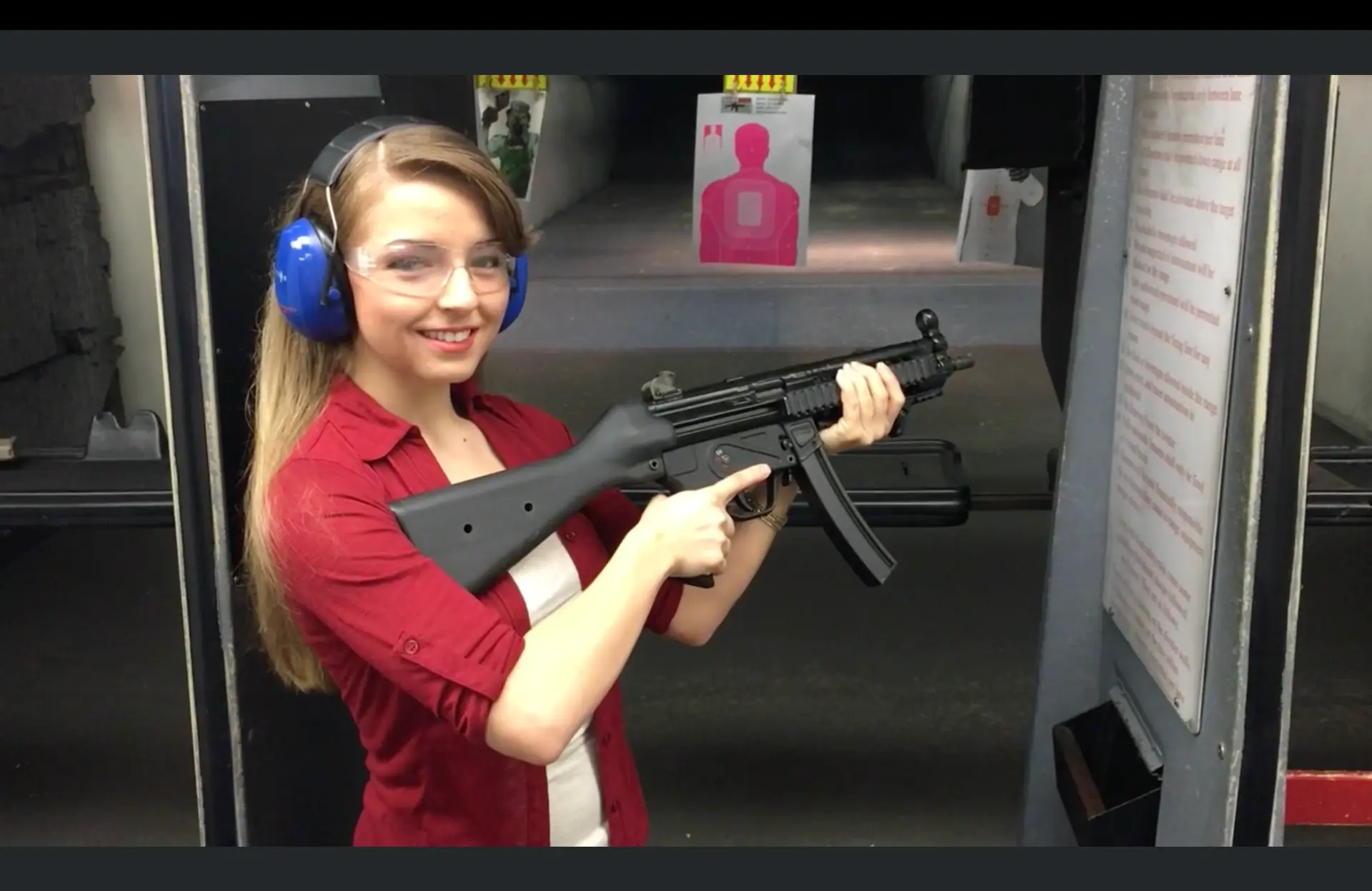 A smiling first-time shooter wearing blue hearing protection and clear eye protection holds an MP5-style submachine gun in an indoor shooting lane
