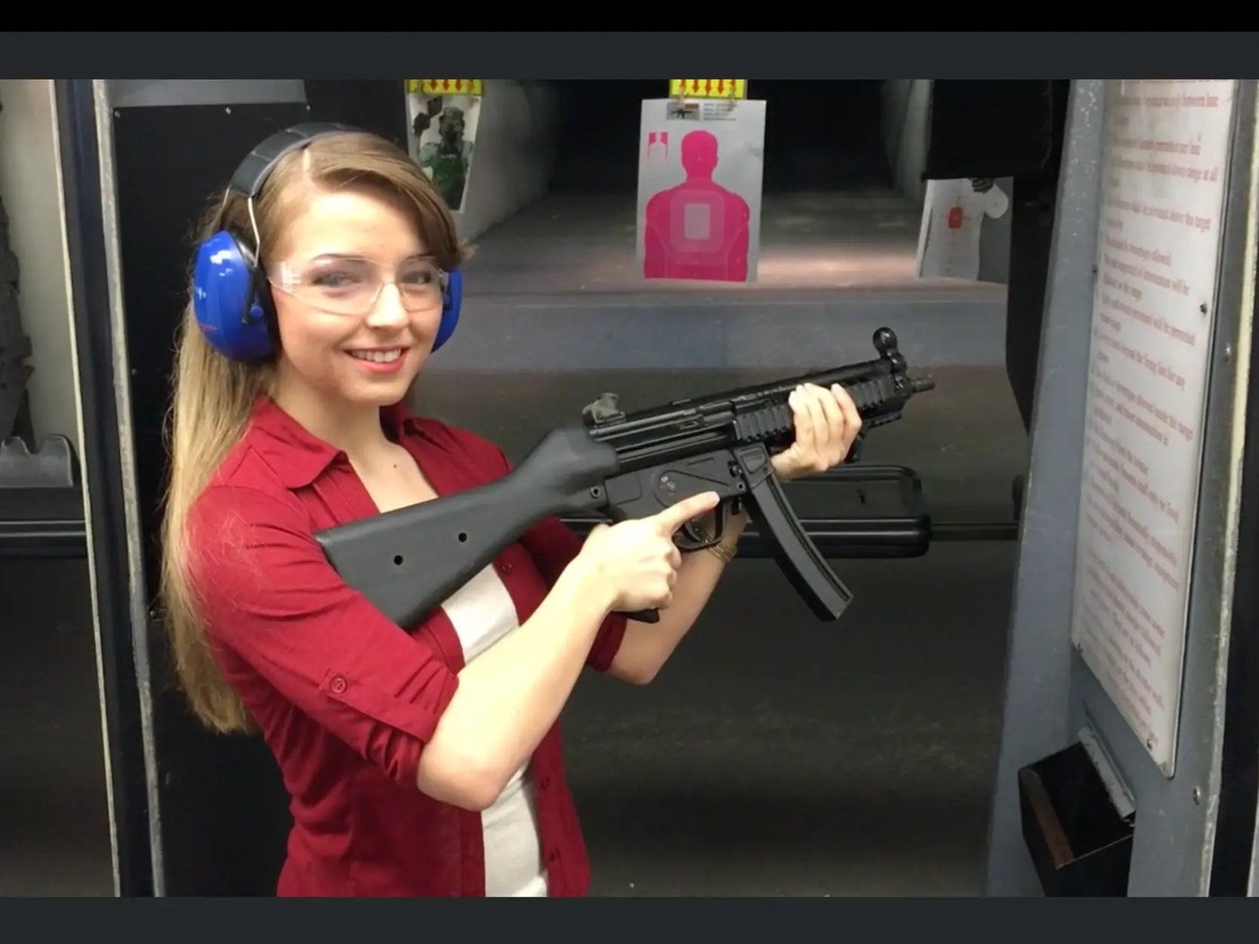 A first-time shooter wearing ear and eye protection holds a submachine gun in an indoor range lane