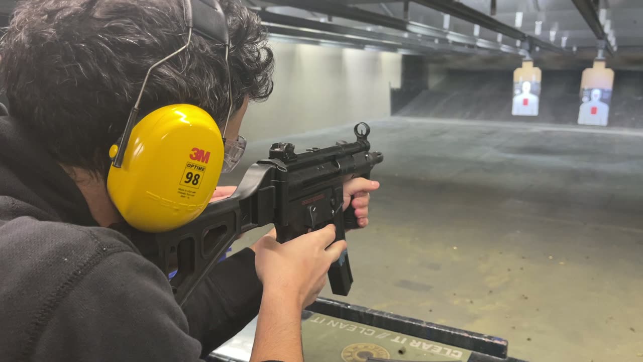 A shooter firing a submachine gun from a bench down an indoor range lane under supervision